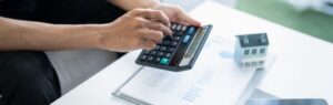 Persona che calcola documenti finanziari con una calcolatrice accanto a un modellino di casa, concetto legato alle tasse sulla casa in Thailandia.