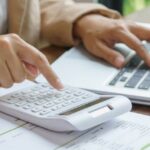 Personne utilisant une calculatrice et un ordinateur portable pour gérer des documents financiers, image liée au thème payer ses impôts en Thaïlande.